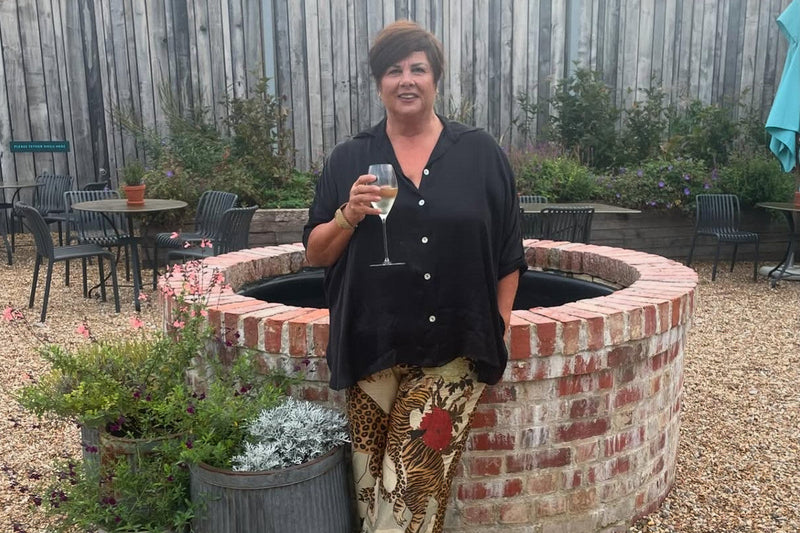 Lyn stands in front of a raised circular pond.  She wears a black shirt and patterned trousers.  She is holding a glass of wine.